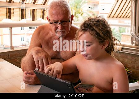 A 70-year-old man and his 9-year-old grandson sitting under a sunny awning on a balcony, focused on exploring a tablet together. Stock Photo