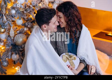 Romantic couple hugging at Christmas tree, wrapped in blanket, exchanging gifts in cozy festive atmosphere by fireplace. Family holiday of New Year in circle of loved ones, happy moment of lovers Stock Photo