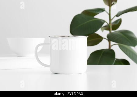Enamel mug mockup with workspace accessories on a white table and a ficus plant. Stock Photo