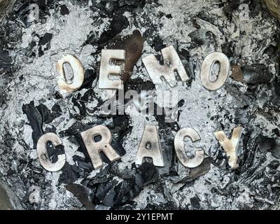 Charred wood letters spelling out the word democracy sit among a pile of ashes in a trash can, suggesting democracy is under attack. Stock Photo