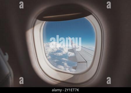 view from the porthole captures a stunning horizon, showcasing the airplane wing against a backdrop of blue skies and fluffy clouds Stock Photo