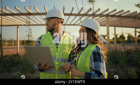 Two Caucasian experienced engineers workers woman female girl man male guy hardhat using laptop pc gadget device computer discuss talking project Stock Photo