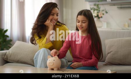 Caucasian family saving money adult woman female mom mother teaching little girl child kid save throwing coins piggy bank financial education Stock Photo