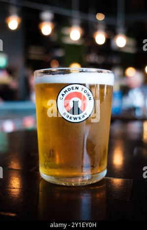 A refreshing glass of craft beer sits on a wooden bar top, featuring the Camden Town Brewery. The vibrant ambiance showcases blurred lights in a bustl Stock Photo