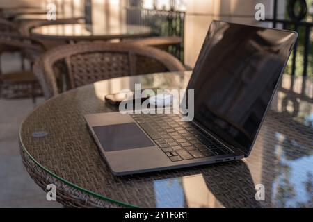A laptop sits open on a table outside, a phone and a wireless mouse beside it. The table is surrounded by wicker chairs. Stock Photo