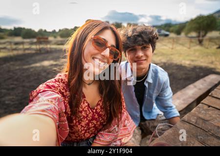 Cheerful young couple taking a selfie in a rustic outdoor setting at sunset. Smiling woman with sunglasses and man sitting on wooden fence, capturing Stock Photo