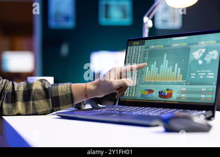 An individual pointing at a screen that shows numbers and financial charts related to stock trading. At a desk, digital laptop is displaying graphs and values for forex trends. Stock Photo