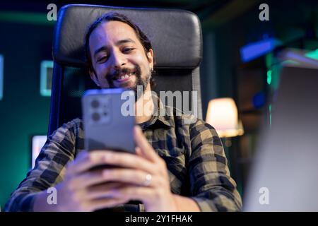 Bearded man sitting in front of laptop holding his cell phone, smiling and browsing the internet. Cheerful young male individual using his mobile device to check his social medias. Stock Photo