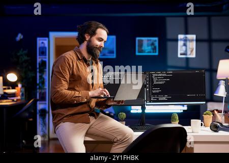 Smiling IT expert doing software quality assurance, reading source code on laptop before implementing it. Happy man sitting on desk inspecting coding on notebook, looking to fix potential issues Stock Photo