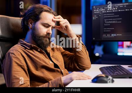 Tired man hurting from too much stress accumulated while doing software quality assurance, reading source code on computer. Developer suffers from headache after inspecting coding on PC for too long Stock Photo