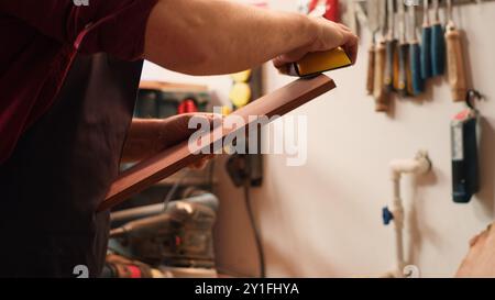 Carpenter doing last touches on piece of wood before using it for furniture assembly, sanding it with sandpaper. Woodworking expert doing woodworking process on lumber block, fixing damages, camera A Stock Photo