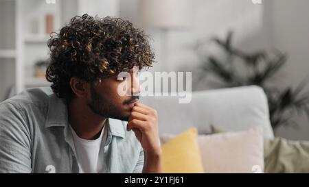 Bored millennial generation man sitting on sofa, feeling lonely at home. Unhappy young male suffering from negative thoughts, having depressive mood Stock Photo