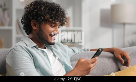 Happy young man with beard chilling and listening his favorite song. Indian brunette man seat on sofa and relax after work, uses smartphone for Stock Photo