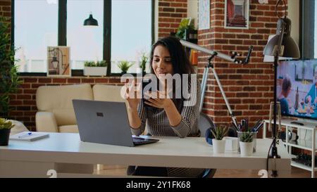 Teleworker checking her phone notifications at home in front of laptop, taking break from work. Woman in cozy apartment office taking pause from job tasks to read messages on smartphone, camera A Stock Photo