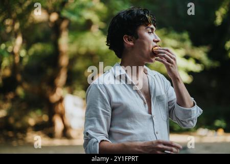 Young man enjoying a snack outdoors in a lush forest environment, wearing a casual white shirt, with sunlight filtering through the trees creating a Stock Photo