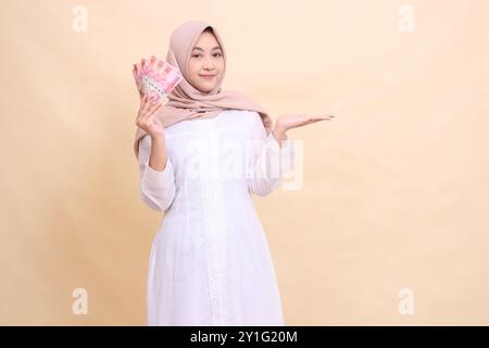 woman indonesia muslim wearing a hijab smiles gracefully holding rupiah cash in her hand and presenting it to the left. Lifestyle, religious, business Stock Photo