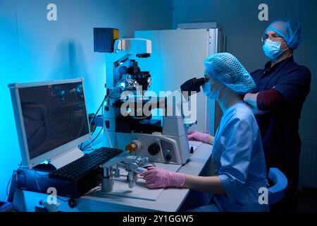 Two lab technicians working with a microscope and computer in a controlled lab environment Stock Photo