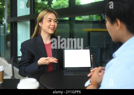 Young businesswoman presenting something on digital tablet to her colleague or client, sitting at outdoor cafe Stock Photo