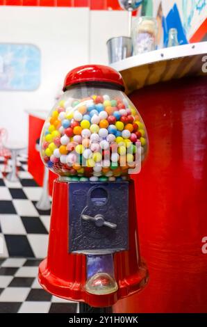 Vintage red gumball machine filled with colorful gumballs standing in a retro diner with black and white checkered floor and red decor. Stock Photo