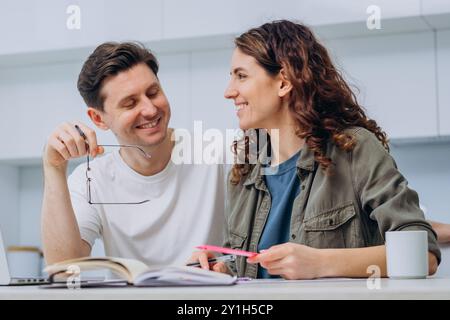 Smiling couple working together at home with books and laptop, enjoying teamwork and shared project, using notebooks and computer in cozy home setting. Family man and woman talking sweetly in kitchen. Stock Photo