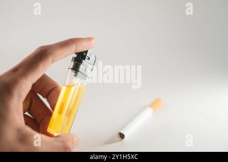 A hand holding a transparent lighter filled with yellow liquid, with a white cigarette lying beside it on a light background. Stock Photo