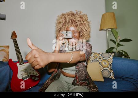 Blonde curly haired black woman using silver camera while taking photo and giving thumb up sign Stock Photo