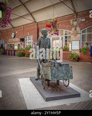 A bronze statue of a barrow boy stands in a railway station foyer.  Flowers are in planters and hanging baskets and a clock is over the booking office Stock Photo