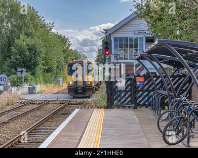 A train approaches a railway station at a road crossing. A signal box is at the end of a platform and there are bicycles in the foreground. Stock Photo