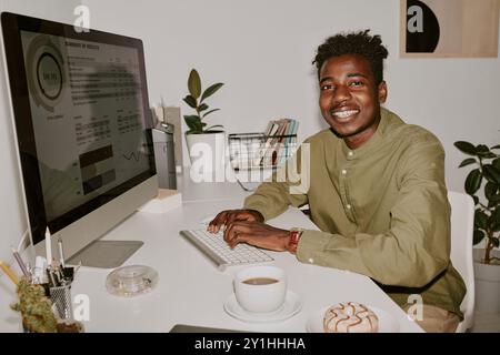 Young male programmer sitting at table and typing on keyboard while posing for portrait in office Stock Photo