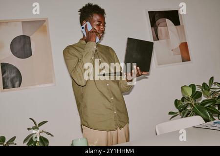 Dutch angle shot of busy young influencer holding laptop while speaking on phone Stock Photo