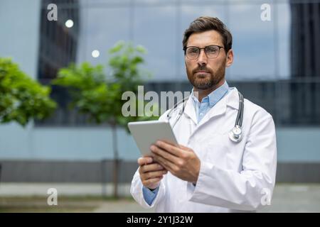 Confident male doctor with stethoscope using tablet outdoors. Healthcare professional focused on digital device for accessing medical data, managing records, and virtual consultations. Stock Photo