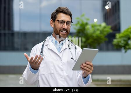 Doctor wearing white coat and stethoscope interacts with patient through video call on tablet. Engages in outdoor telemedicine session in urban area, showcasing modern healthcare technology. Stock Photo