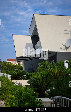 Modern white cubic house with many edges is standing under the blue sky, surrounded by green plants. Stock Photo