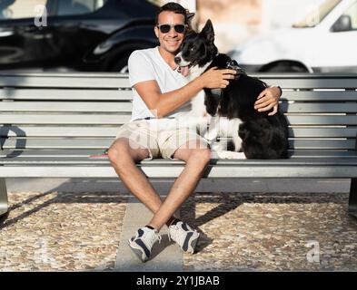 A man sits on a bench with his dog. The man is smiling and the dog is looking up at him. The scene is peaceful and happy Stock Photo