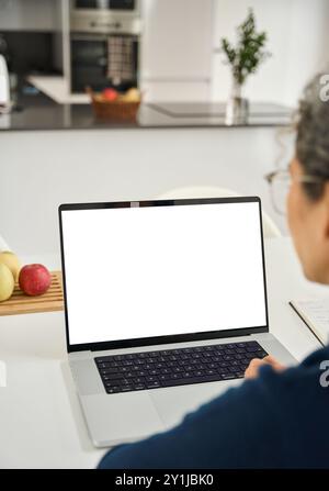 Mature woman looking at white mock up laptop screen using laptop at home. Stock Photo