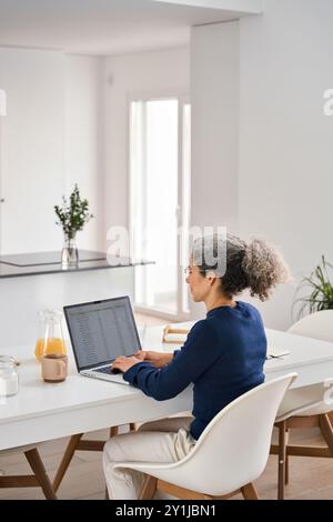 Busy middle aged woman sitting at table and using laptop at home kitchen. Stock Photo