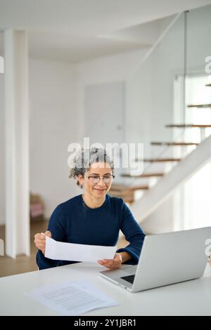 Mature woman looking at laptop holding documents hybrid working at home. Stock Photo