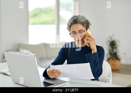 Busy middle aged woman working from home making phone call checking document. Stock Photo