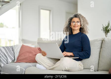 Happy middle aged woman using laptop computer sitting on sofa at home. Stock Photo