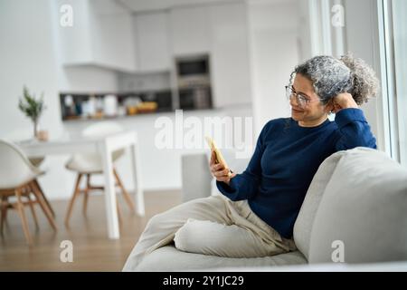 Happy middle aged woman using mobile phone messaging relaxing on couch at home. Stock Photo