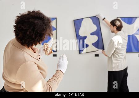 Back view of two art gallery workers hanging paintings on wall and giving directions while planning exhibition copy space Stock Photo