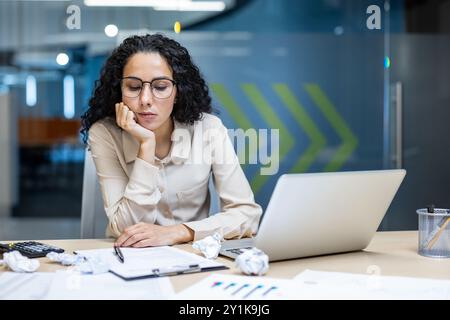 Hispanic businesswoman looking exhausted working on laptop in office. Surrounded by crumpled papers and documents, expressing stress and fatigue. Concept of overwork, deadline pressure Stock Photo