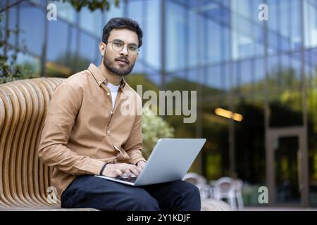 Confident young man wearing glasses using laptop on wooden bench in outdoor urban area. Casual attire, focused expression, reflecting modern lifestyle and digital connectivity in professional setting Stock Photo