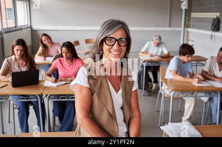 Portrait of a mature teacher at work looks at camera smiling while students study in the background. Stock Photo