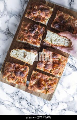 A hand picking a slice of freshly baked focaccia bread from a wooden board. The bread is golden, topped with rosemary sprigs and salt flakes and has a Stock Photo