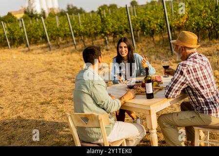 Three friends gather around a rustic table in a vineyard, enjoying wine, fresh fruits, and each other's company on a sunny day filled with joy and lau Stock Photo