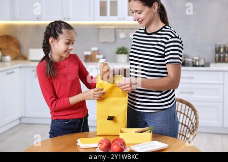 Mother with her daughter putting bottle of drink into lunch bag at wooden table in kitchen. Preparing for school Stock Photo