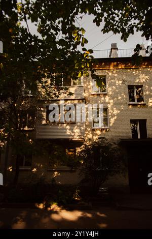 On a summer evening, the warm and cozy light of the sun shines through the leaves of the trees, casting a gentle glow on the apartment building. Stock Photo