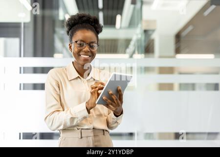 Smiling African American businesswoman holding tablet in contemporary office. Represents success, technology, and confidence in digital work environment. Ideal for themes of business, leadership, and professional growth. Stock Photo