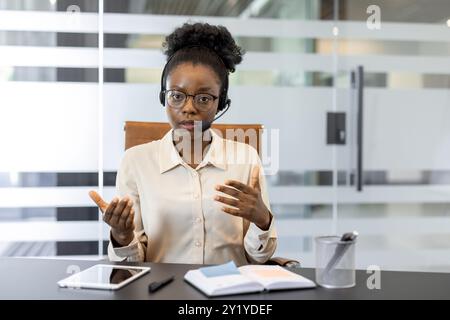 Confident African American businesswoman wearing headset engaged in video conference in office. Professional discussion with gestures, utilizing tablet and notebook for efficient communication Stock Photo